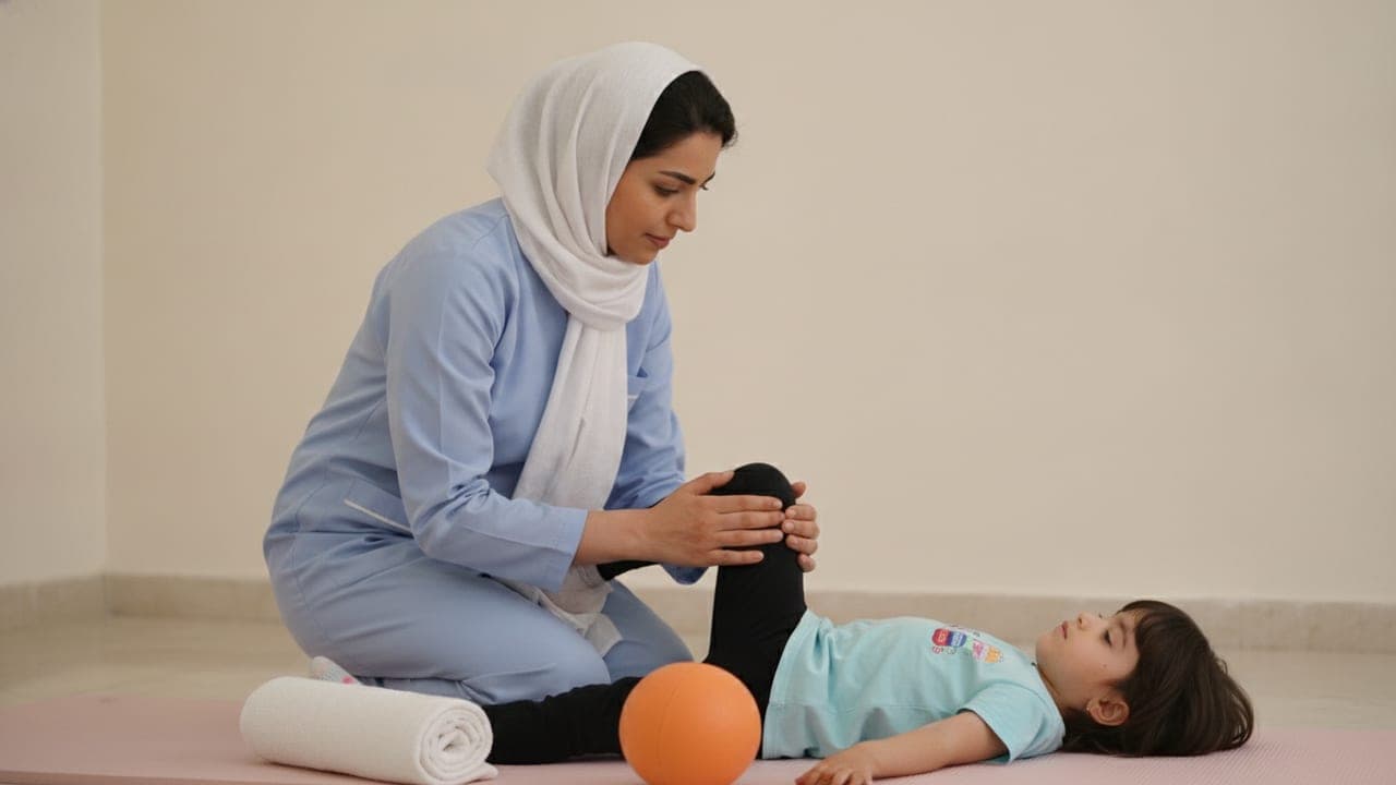 پرستار در منزل روی تشک ورزشی با دختربچهی مبتلا به فلج مغزی تمرین کششی آرام زانو و پا انجام میدهد